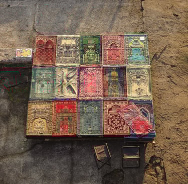 An aerial view of 16 prayer rugs arranged in a grid in an outdoor concrete setting.