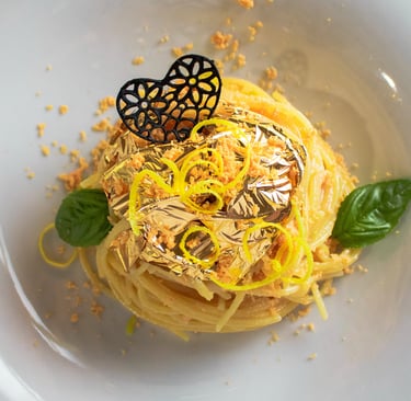 Spaghetti con foglia d'oro, cuore nero traforato, briciole arancioni e basilico in piatto bianco