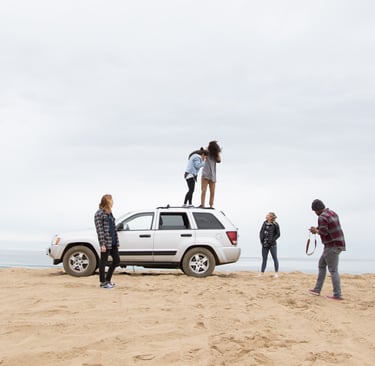 A group of friend near a beach front with a car parked in the middle enjoying their day and giggling