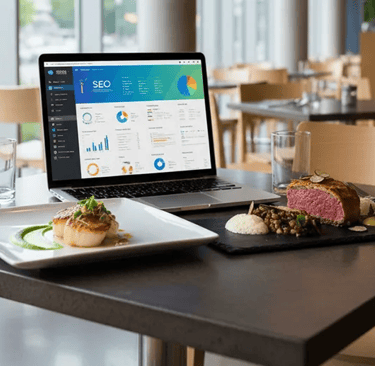 Mesa de restaurante com pratos gourmet e notebook exibindo gráficos de SEO, representando o marketing digital gastronômico.