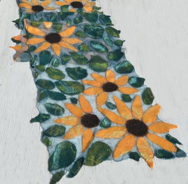 Handmade nuno felted scarf featuring yellow sunflowers and green leaves on a white wooden surface.