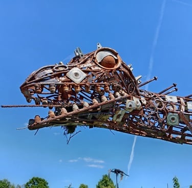 A rustic metal dinosaur sculpture made from scrap metal and recycled parts against a blue sky.