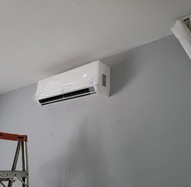A wall-mounted ductless mini split air conditioner unit being installed on a grey wall with a stepladder nearby.