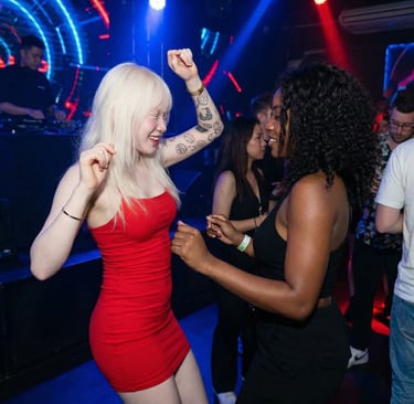 Two diverse women dancing at a lively nightclub with neon lights and a DJ performing in the background.