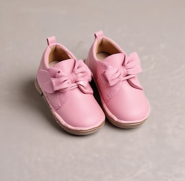 A pair of delicate pink baby shoes with lacy bows and pearl embellishments are placed on a soft surface next to a pink pacifier adorned with rhinestones. Behind them, a glass jar contains a bouquet of artificial roses in various colors, including deep red, pastel pink, and cream, with green leaves.