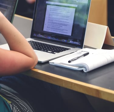 Someone's elbow rests on a table next to an open notebook and laptop.