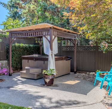 A backyard hot tub under a wooden gazebo with blue Adirondack chairs on a stone patio.
