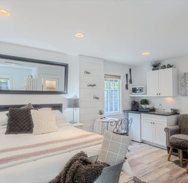 Modern studio apartment featuring a white bed, large wall mirror, and compact kitchenette with white cabinets.