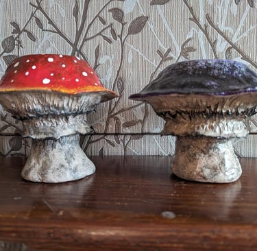 Hand-painted red amanita and purple clay mushroom figurines on a wooden shelf.
