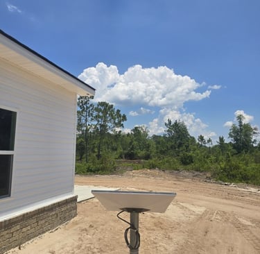 A rectangular Starlink satellite dish mounted on a pole near a rural white house under a bright blue sky.