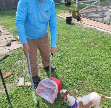 Robert using a sanitized scooper to remove waste from a customers back yard, small dog watching him