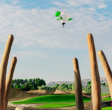 A green dinosaur hot air balloon flies over a modern asphalt pump track with wooden sculptures.