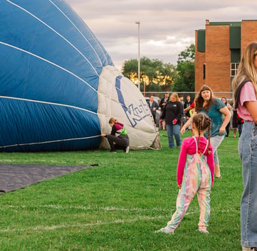 Spectators watch a large blue hot air balloon being inflated on a green field during a community festival.