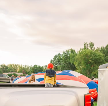 A child in a pickup truck watching a colorful hot air balloon inflate at a festival.
