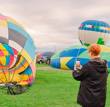 A woman takes a photo of colorful hot air balloons inflating on a green field during a festival.