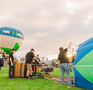 A flight crew prepares colorful hot air balloons for takeoff in a grassy field during a festival.