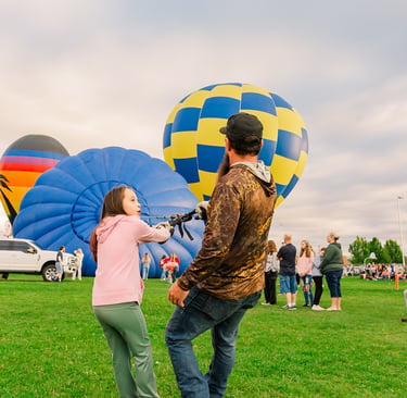 A young girl and man pull ropes to inflate colorful hot air balloons at an outdoor festival.