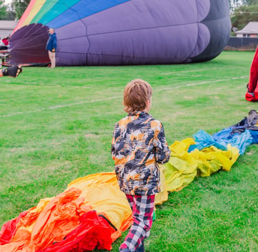 A young boy watches a colorful rainbow hot air balloon being inflated on a green grass field.