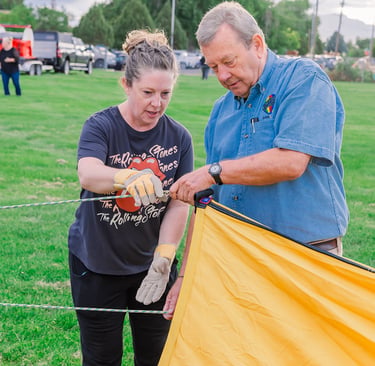 A man and woman prepare a yellow hot air balloon envelope for flight in a grassy field.