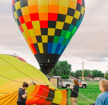 A vibrant rainbow checkered hot air balloon inflates on a grassy field with a ground crew.