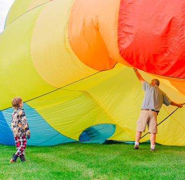 A child watches as a man prepares a colorful rainbow hot air balloon for inflation on a grassy field.