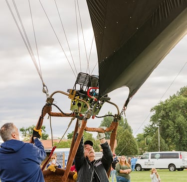 Ground crew preparing a hot air balloon basket and burner for flight at an outdoor festival.