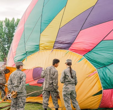 Military cadets in camouflage uniforms preparing a colorful hot air balloon for flight on a grassy field.