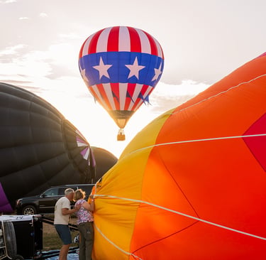 A vibrant patriotic hot air balloon takes flight as crews inflate more balloons during a sunset festival.