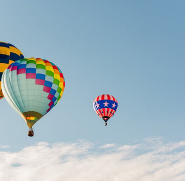 Three colorful hot air balloons floating in a clear blue sky with light clouds during a festival.