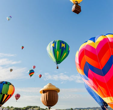 Colorful hot air balloons in various shapes and sizes take flight over a grassy field during a festival.