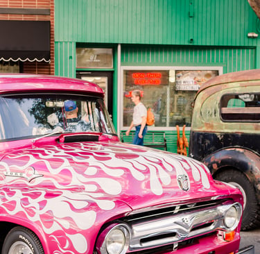 Vintage pink Ford F100 truck with white flame decals parked on a city street near storefronts.