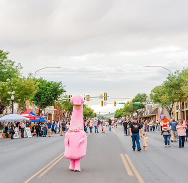 A large pink dinosaur mascot walks down a crowded city street during a summer community festival.