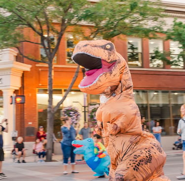 A person wearing a funny inflatable T-Rex dinosaur costume running down a city street during a parade.
