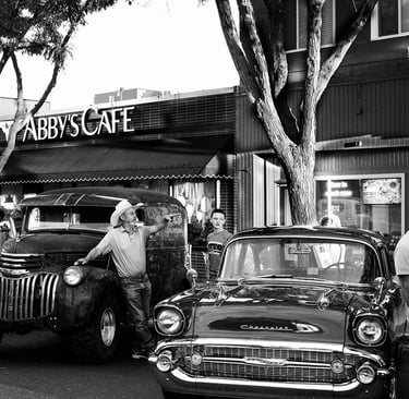 Black and white photo of a cowboy with a vintage Chevrolet car and truck outside Abby's Cafe.