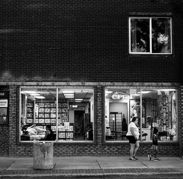 Black and white street photography of people walking past a brick tattoo parlor storefront window.