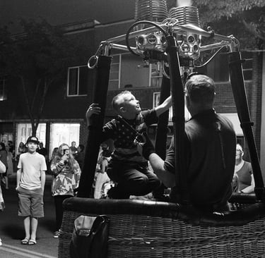 A young boy sits in a wicker hot air balloon basket at night as a pilot ignites the gas burner.