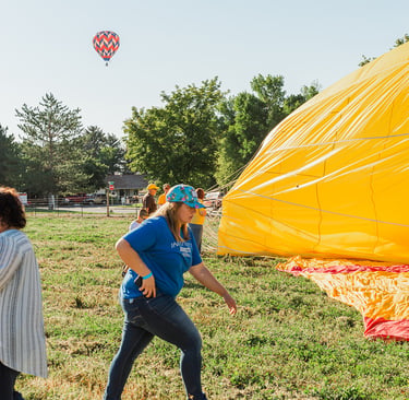 Volunteers prepare a large yellow hot air balloon for flight in a grassy field on a sunny day.