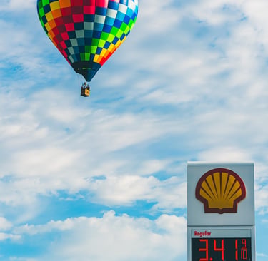 A colorful checkered hot air balloon floats in a blue sky above a Shell gas station price sign.