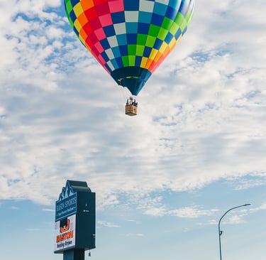 A colorful rainbow checkered hot air balloon flying over a city street with a commercial sign.