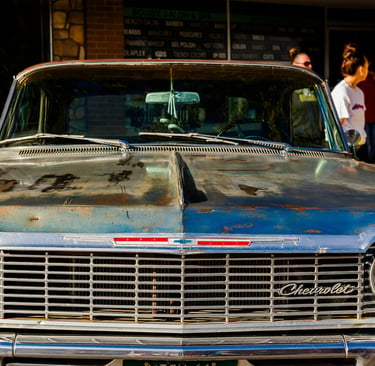 Front view of a classic blue Chevrolet Impala with a rusted hood parked in front of a local salon.