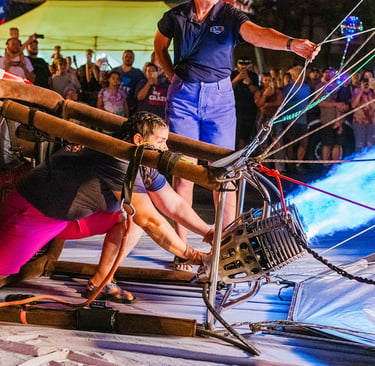 A crew member operates a propane burner with a blue flame to inflate a hot air balloon basket at night.