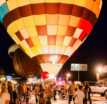 Crowd gathers for a colorful hot air balloon glow event at night with illuminated checkered balloons.