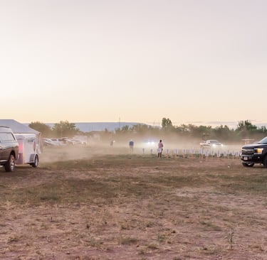 Trucks and trailers parked in a dusty field during an outdoor sunset event with people gathered.