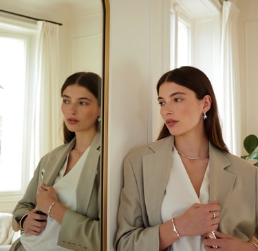 Elegante Frau mit minimalistischem Silberschmuck – Kette, Ohrringe, Armband – steht vor Spiegel im modernen Raum