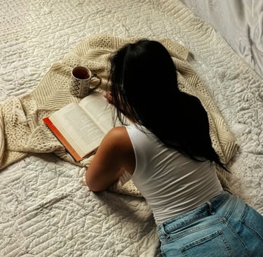 A woman relaxes on a cozy bed with a book, coffee, and a textured knit blanket.