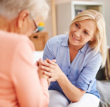 A blonde female caregiver provides compassionate home care while holding hands with an elderly woman.