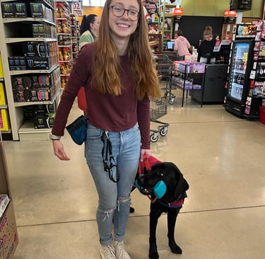 a student dog trainer smiles proudly as her dog holds an object in the grocery store