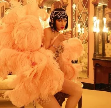 Taylor vestindo penas cor-de-rosa, com um copo de champanhe, em um quarto luxuoso.