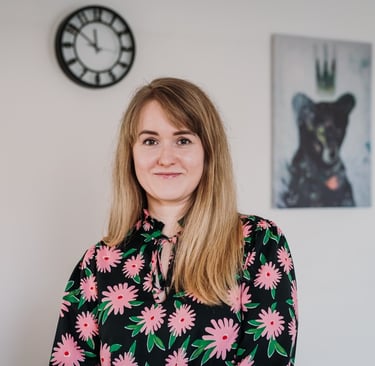 Psichoterapeutė Evelina A. stovinti šviesiame kambaryje; fone matosi sieninis laikrodis, paveikslas