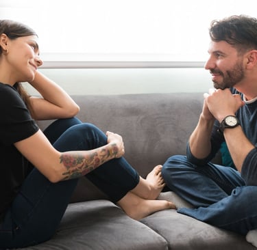 Moteris ir vyras sėdi ant minkštos sofos ir šypsosi žiūrėdami vienas į kitą.